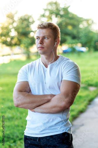 The man atlen stands in the park with headphones, a white T-shirt. Summer lifestyle, motivation is strong. Muscular arms.