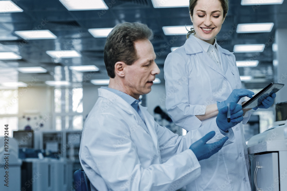 Professional scientists. Delighted smart positive woman holding a table and looking at her colleague while being happy about his success