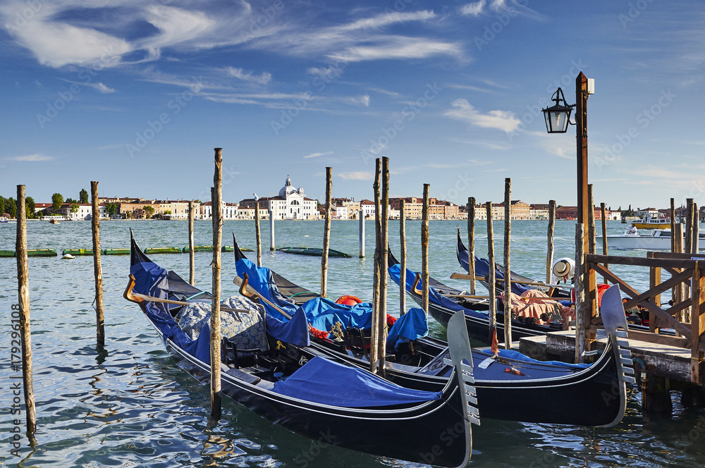 Fototapeta premium Blue gondolas in Venice moored to the jetty between wooden piles with view of the sea and the palace at sunset