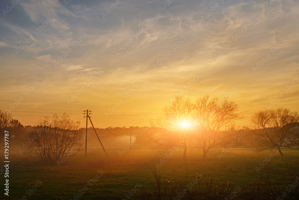 Obraz premium An landscape of autumn and foggy countryside greenfield with village on a background at sunset