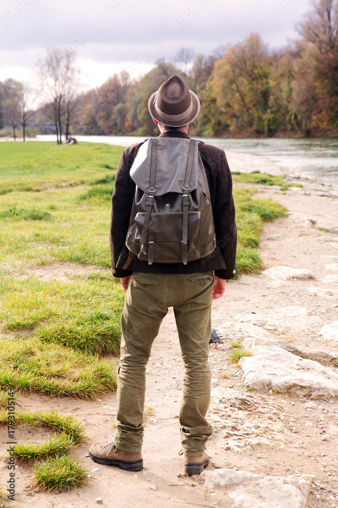 backside of bavarian man with backpack and standing by river backside of bavarian man with backpack and standing by river