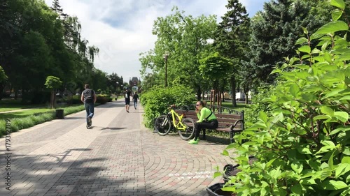 Pretty girl cyclist bicycle using smartphone park sunny day cute city parks active young woman cyclist cycling talking phone alley downtown guy rides scooter passers green grass carpet lawn flowerbeds