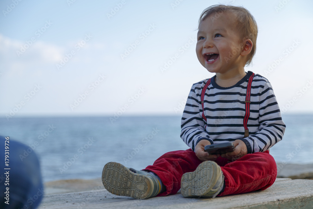 Fototapeta premium A baby boy is playing with mobile phone on a beach