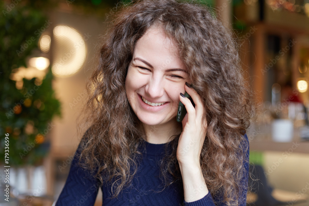 Cheerful excited young European female with curly gorgeous hair wearing blue sweater laughing with tender as she receives good positive news during phone conversation with friend. Lifestyle concept. Cheerful excited young European female with curly gorgeous hair wearing blue sweater laughing with tender as she receives good positive news during phone conversation with friend. Lifestyle concept.