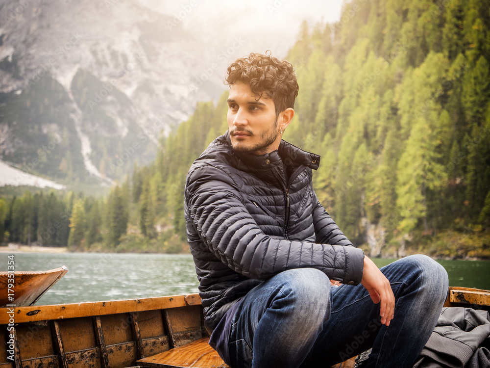 Fototapeta premium Handsome tattooed man sitting in boat on background of woods on shore of lake looking away.