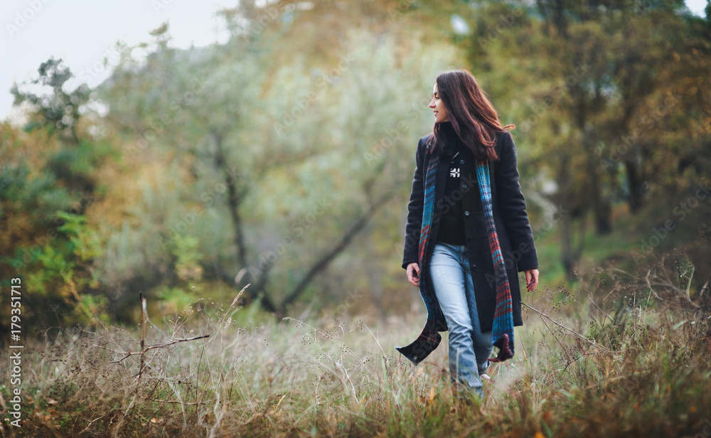 A pretty girl in a woolen coat with a long scarf walks through the autumn forest against the backdrop of nature.