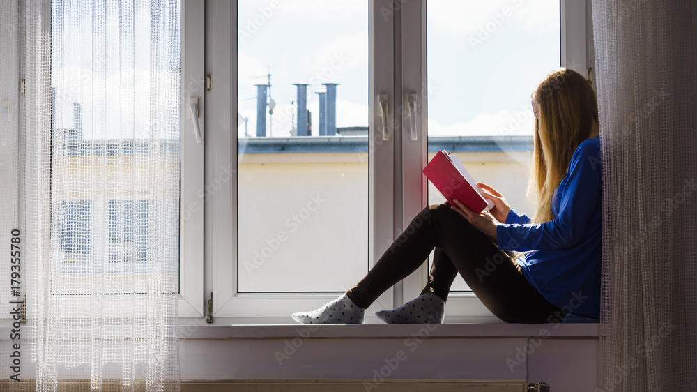 Woman sitting on window sill reading book at home Woman sitting on window sill reading book at home