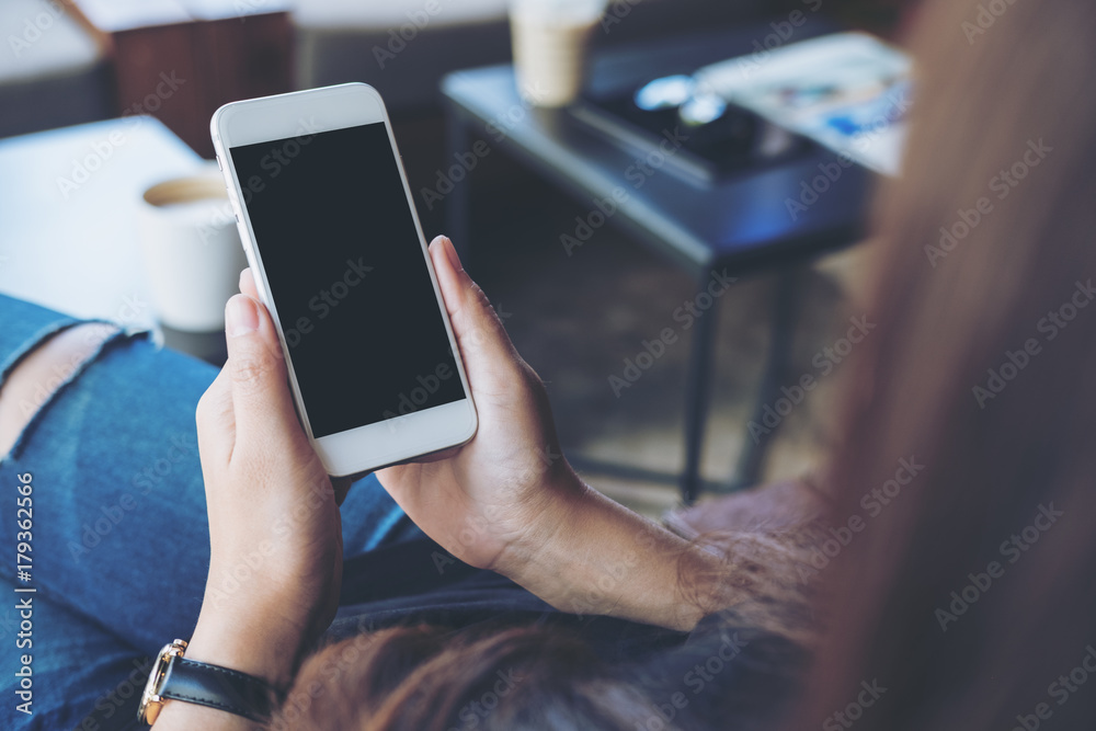 Mockup image of woman's hands holding white mobile phone with blank black screen on thigh and coffee cup on the table