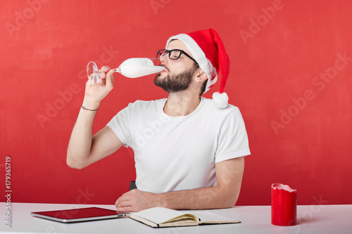 Cheers! Man drinks at work. Young attractive hipster in Christmas hat celebrating New Year at work place, drinking milk from the glass party glass, spending time with gadgets and devices indoors. photo