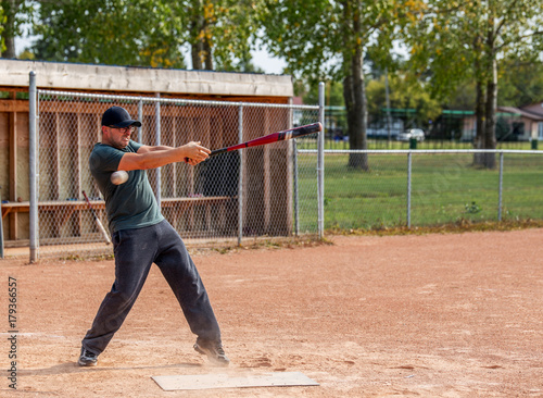 A man in his thirties in sweat pants and t-shirt swinging and missing a baseball with lips curled in effort at a summer time baseball diamond