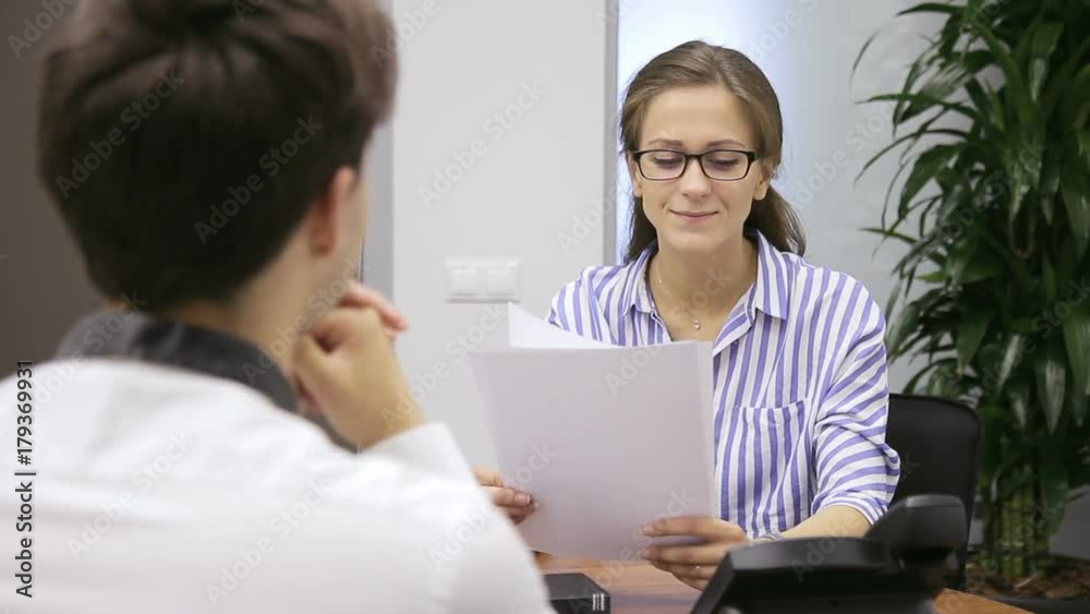 Business woman in glasses holds an interview with competitor in office. Portrait of lady in striped blouse who holds documents in her hands and communicates well with man opposite. Business woman in glasses holds an interview with competitor in office. Portrait of lady in striped blouse who holds documents in her hands and communicates well with man opposite.