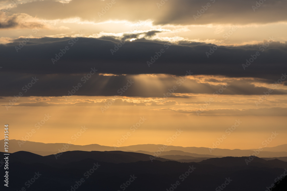A silhouette of a mountain peak at sunset, beneath sun rays coming out through some clouds