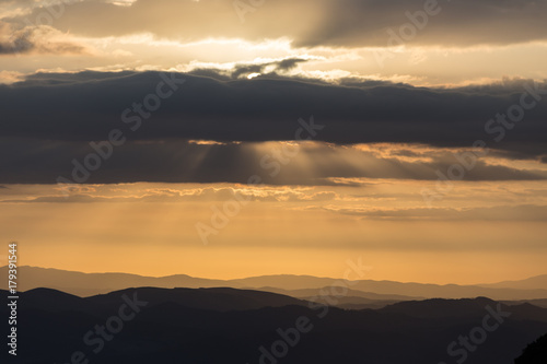 A silhouette of a mountain peak at sunset, beneath sun rays coming out through some clouds © Massimo