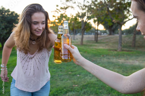 Two young women joking on lawn in park and toasting with beer bottles having fun.  