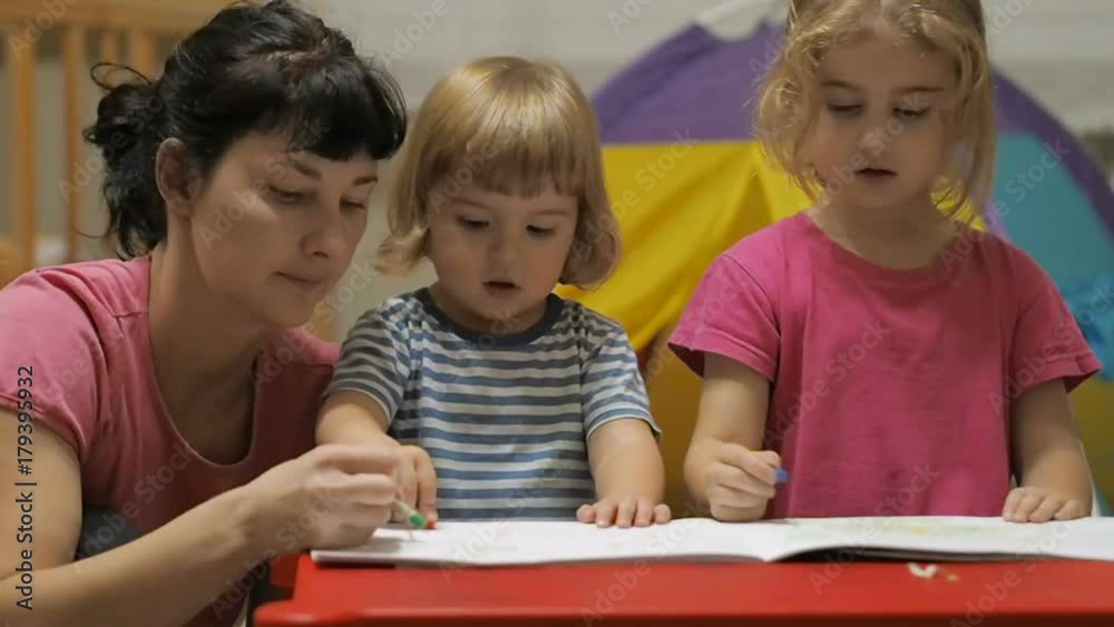 The educator and two little children girl and boy sitting at table drawing. Brother and sister and their mother drawing with colorful crayons and markers The educator and two little children girl and boy sitting at table drawing. Brother and sister and their mother drawing with colorful crayons and markers