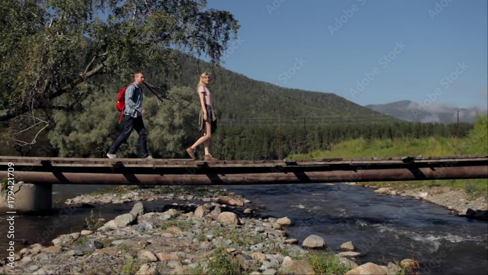 couple of travelers with a backpack walk on a bridge across a mountain river couple of travelers with a backpack walk on a bridge across a mountain river