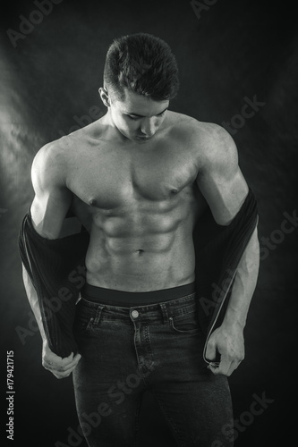 Handsome young muscular man shirtless wearing jeans and sleeveless vest, on black background in studio shot