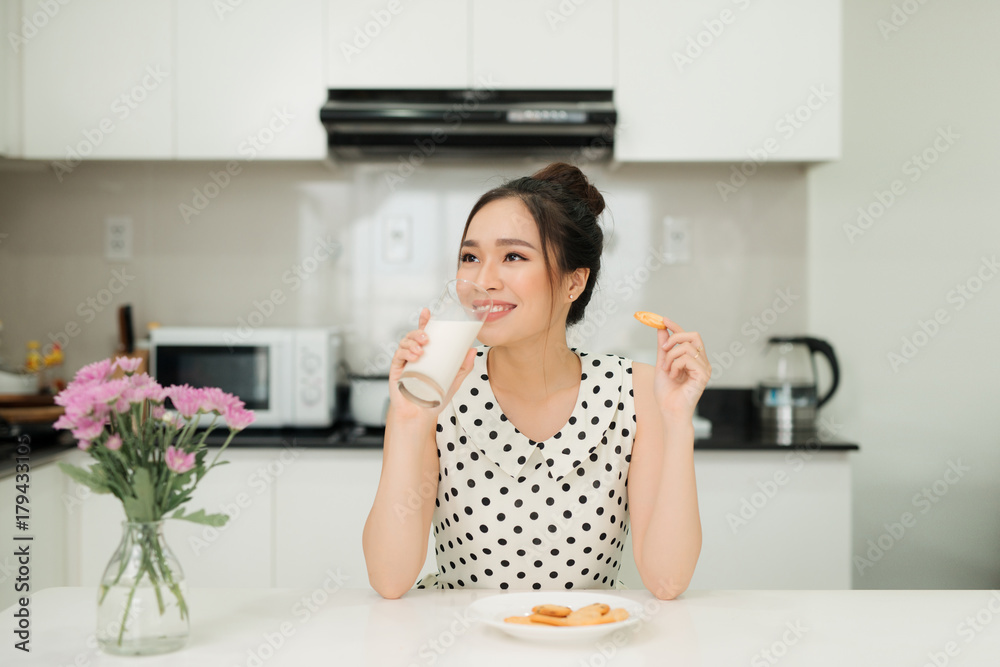 Young asian woman holding milk glass bite cookie in her kitchen Young asian woman holding milk glass bite cookie in her kitchen