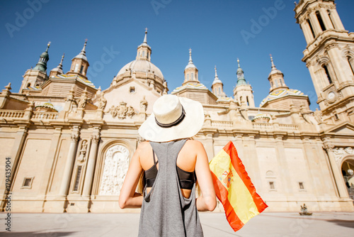 Young woman tourist standing back with spanish flag in front of the famous cathedral on the central square during the sunny weather in Zaragoza city, Spain