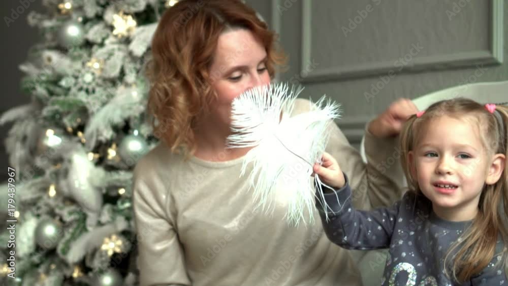 Mother with the daughter at a Christmas tree sit on a sofa and laugh. Mother tickles the fair-haired daughter a white feather. Family around christmas tree. Slow mothion Mother with the daughter at a Christmas tree sit on a sofa and laugh. Mother tickles the fair-haired daughter a white feather. Family around christmas tree. Slow mothion
