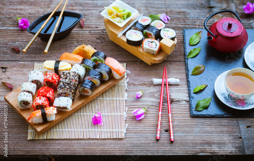 Red teapot, green leaves, slate tray and red chopsticks on old wooden table, Sushi rolls, nigiri, raw salmon steak, rice, cream cheese, avocado, lime, pickled ginger, asian background, top view, 