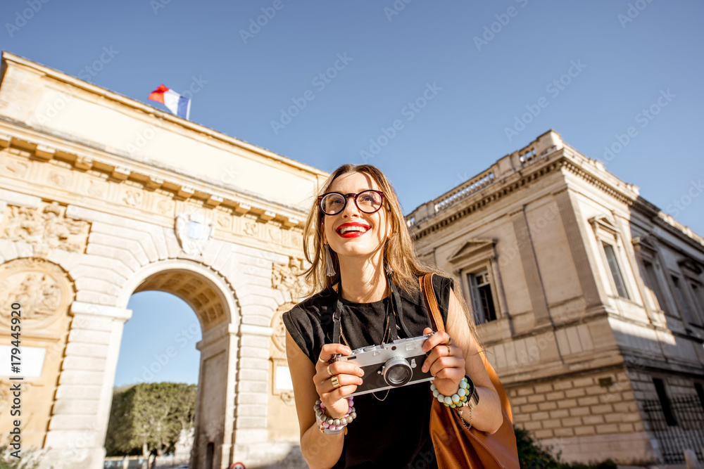Fototapeta premium Portrait of a young happy woman tourist with photo camera in front of the famous Triumphal Arch during the morning light in Montpellier city, France