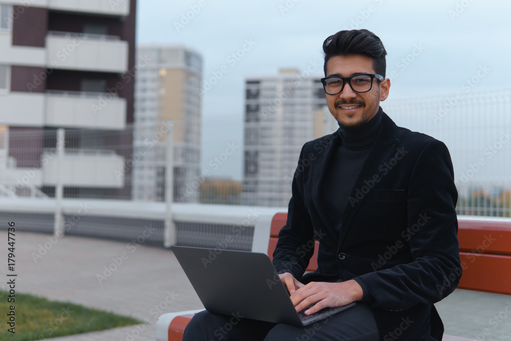 Young businessman in eyeglasses work on the laptop sitting outdoors. Young businessman in eyeglasses work on the laptop sitting outdoors.