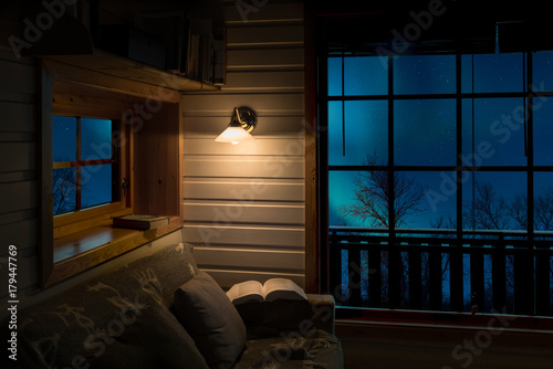 Still life in a cozy cottage with open book illuminated by lamp and northern light outside the windows.