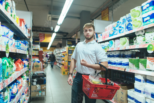 A man with a product basket strolls around the supermarket and chooses goods. A man buys food in a supermarket.