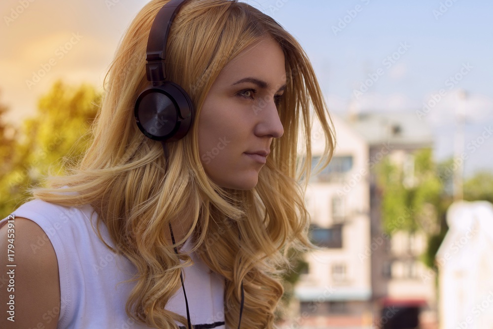 Young beautiful woman with blond hair listens to music in summer during sunset