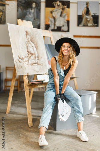 Female artist wears black hat posing near picture indoor the studio