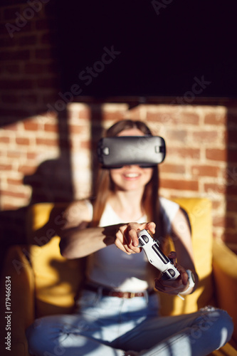 Girl wears white shirt enjoying virtual reality head set indoor sitting on the yellow armchair before bricks wall