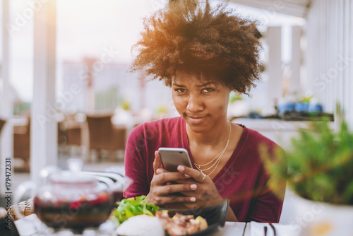 Young Brazilian teenage girl with afro hair is suspiciously looking at the camera while sitting in street cafe outdoors and taking pictures of fresh salad and her other food using smartphone camera