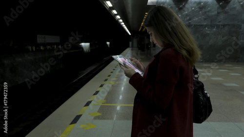 Girl in red coat using smartphone or tablet at subway station and waits for the train. Woman use of cellphone and standing at city subway staton. photo