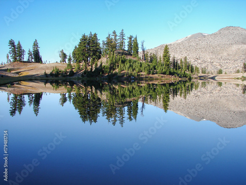A small group of trees reflect off the smooth surface of a clear blue lake at tree line high in the mountains.