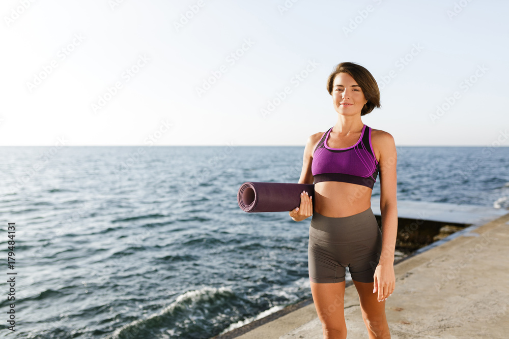 Portrait of young smiling woman with dark short hair holding yoga mat in hand while walking by the sea. Nice lady in sporty top and shorts standing and happily looking in camera with sea on background Portrait of young smiling woman with dark short hair holding yoga mat in hand while walking by the sea. Nice lady in sporty top and shorts standing and happily looking in camera with sea on background