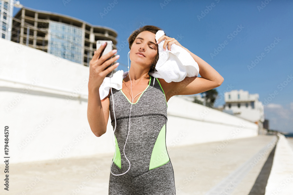 Portrait of beautiful woman with brown short hair in modern gray sport suit standing with towel on shoulders and cellphone in hands while dreamily looking in it isolated