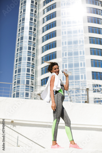 Portrait of joyful woman in modern gray sport suit and pink sneakers standing with white towel on shoulder and happily looking in camera with high building on background