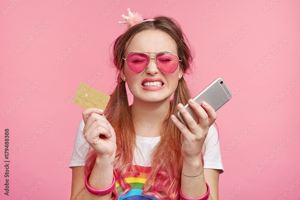Horizontal portrait of upset miserable woman wears trendy sunglasses, holds plastic card and mobile phone, being short of money, can`t buy new clothes or cosmetics, has bad mood. Online shopping Horizontal portrait of upset miserable woman wears trendy sunglasses, holds plastic card and mobile phone, being short of money, can`t buy new clothes or cosmetics, has bad mood. Online shopping