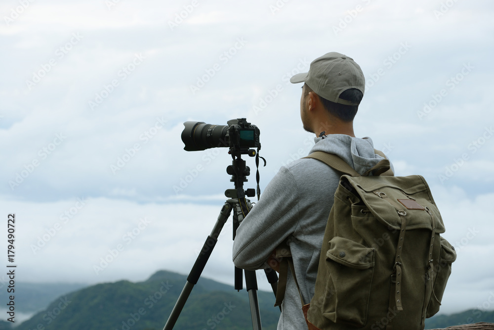 Young tourist man with backpack looking through camera at the sea of fog