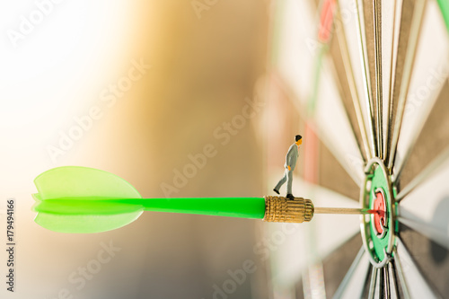 Miniature people: Small business man on red dart arrow hitting in the target center of dartboard with modern city and sunset background. Target business, achieve and victory concept .