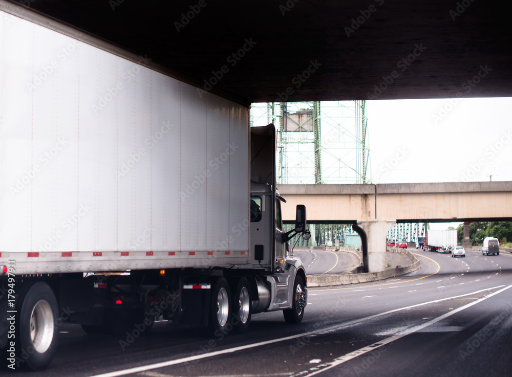 White big rig semi truck with day cab and dry van trailer running on the road under bridge White big rig semi truck with day cab and dry van trailer running on the road under bridge