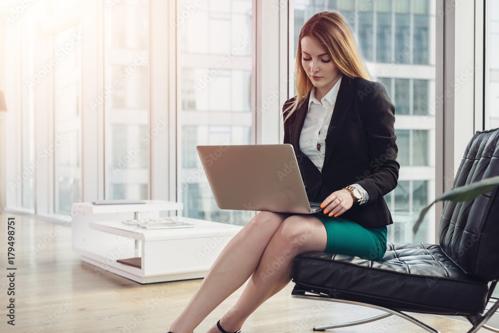 Female chief economist analyzing data using laptop sitting on armchair in modern office Female chief economist analyzing data using laptop sitting on armchair in modern office