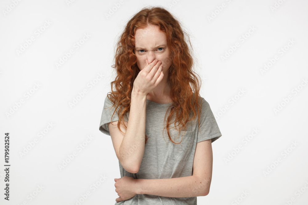 Closeup headshot of young attractive European woman with red hair isolated on white background holding her nose with wry face showing deep dissatisfaction and disgust with unpleasant smell or facts Closeup headshot of young attractive European woman with red hair isolated on white background holding her nose with wry face showing deep dissatisfaction and disgust with unpleasant smell or facts