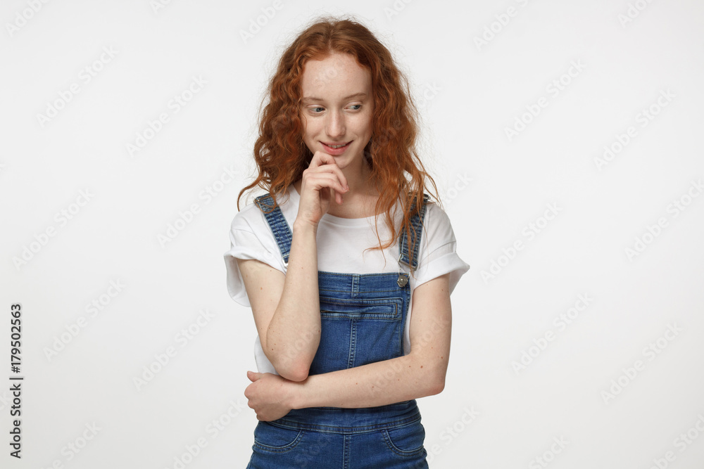 Horizontal portrait of young attractive European woman with red hair isolated on white background standing with arms crossed, touching her chin with fingers, looking down with shy interested smile