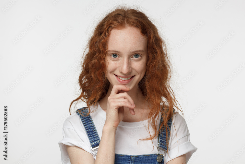 Indoor portrait of young teenage girl with red curly hair isolated on white background looking at camera with joyful smile pressing fingers to her chin as if analyzing and choosing from alternatives Indoor portrait of young teenage girl with red curly hair isolated on white background looking at camera with joyful smile pressing fingers to her chin as if analyzing and choosing from alternatives