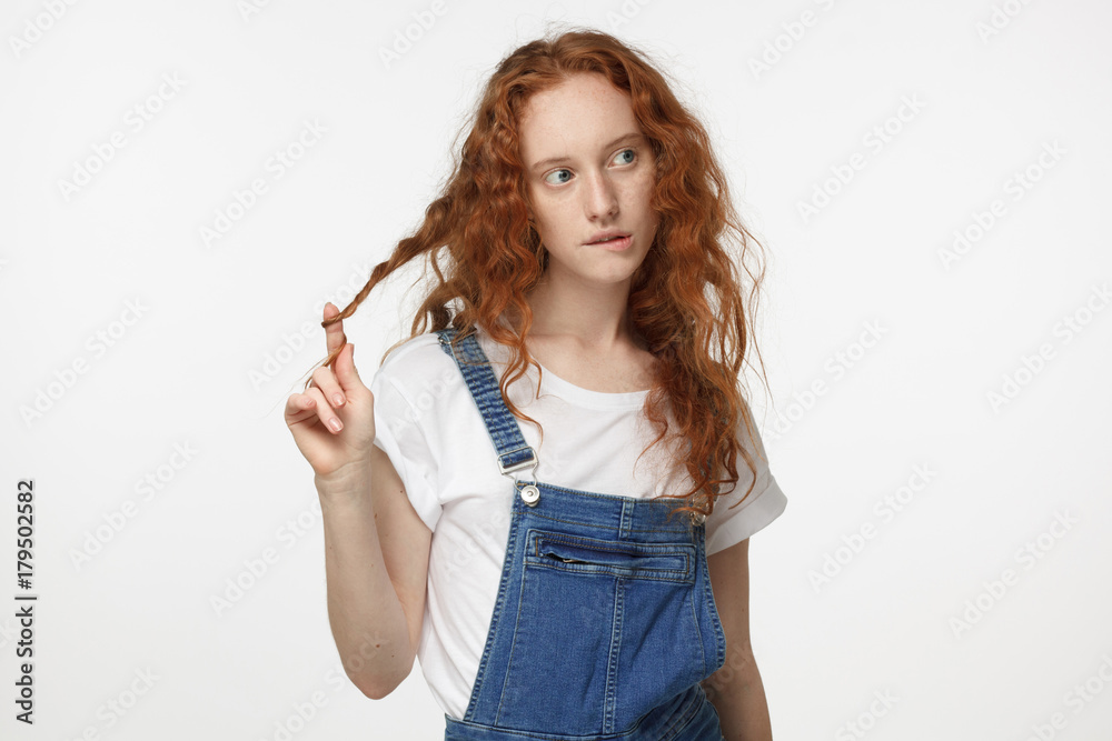 Closeup photo of young teenage girl with red curly hair isolated on white background wearing white T-shirt and denim jumpsuit biting her lip and twisting her hair looking bewildered making decision Closeup photo of young teenage girl with red curly hair isolated on white background wearing white T-shirt and denim jumpsuit biting her lip and twisting her hair looking bewildered making decision