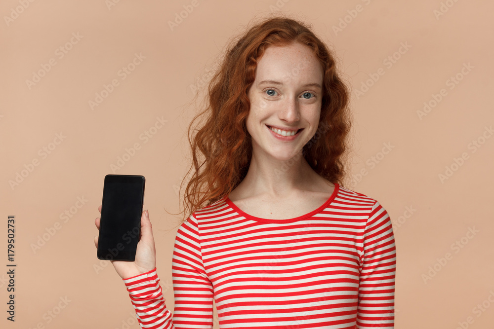 Closeup of young smiling pretty curly redhead girl in striped t-shirt, holding and showing blank smartphone with copyspace for advertisement of applications, websites, goods and services Closeup of young smiling pretty curly redhead girl in striped t-shirt, holding and showing blank smartphone with copyspace for advertisement of applications, websites, goods and services
