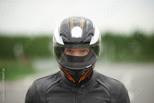 Close up portrait of confident handsome young motorbiker wearing stylish helmet and leather jacket staring at camera with eyes full of determination, ready for motor race on summer day outdoors © Anatoliy Karlyuk