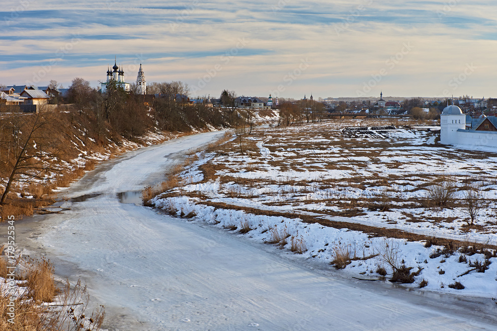 Early spring in Suzdal/City landscape of Suzdal. River, ice-bound, In the flood plain lies snow. On the slope of the hill, the snow melted and last year's grass is visible.Suzdal.Golden Ring of Russia